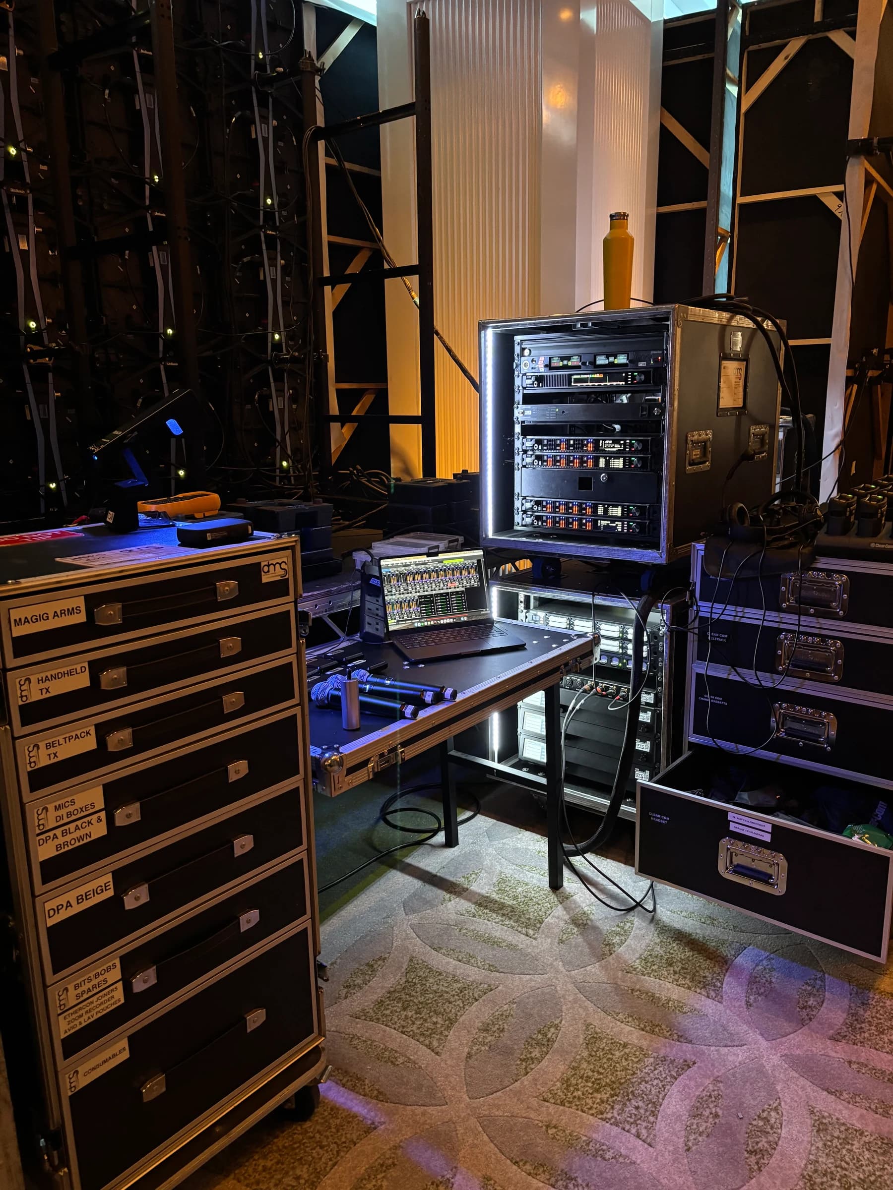 Backstage equipment racks, a laptop and handheld microphones surrounded by flight cases and cabling