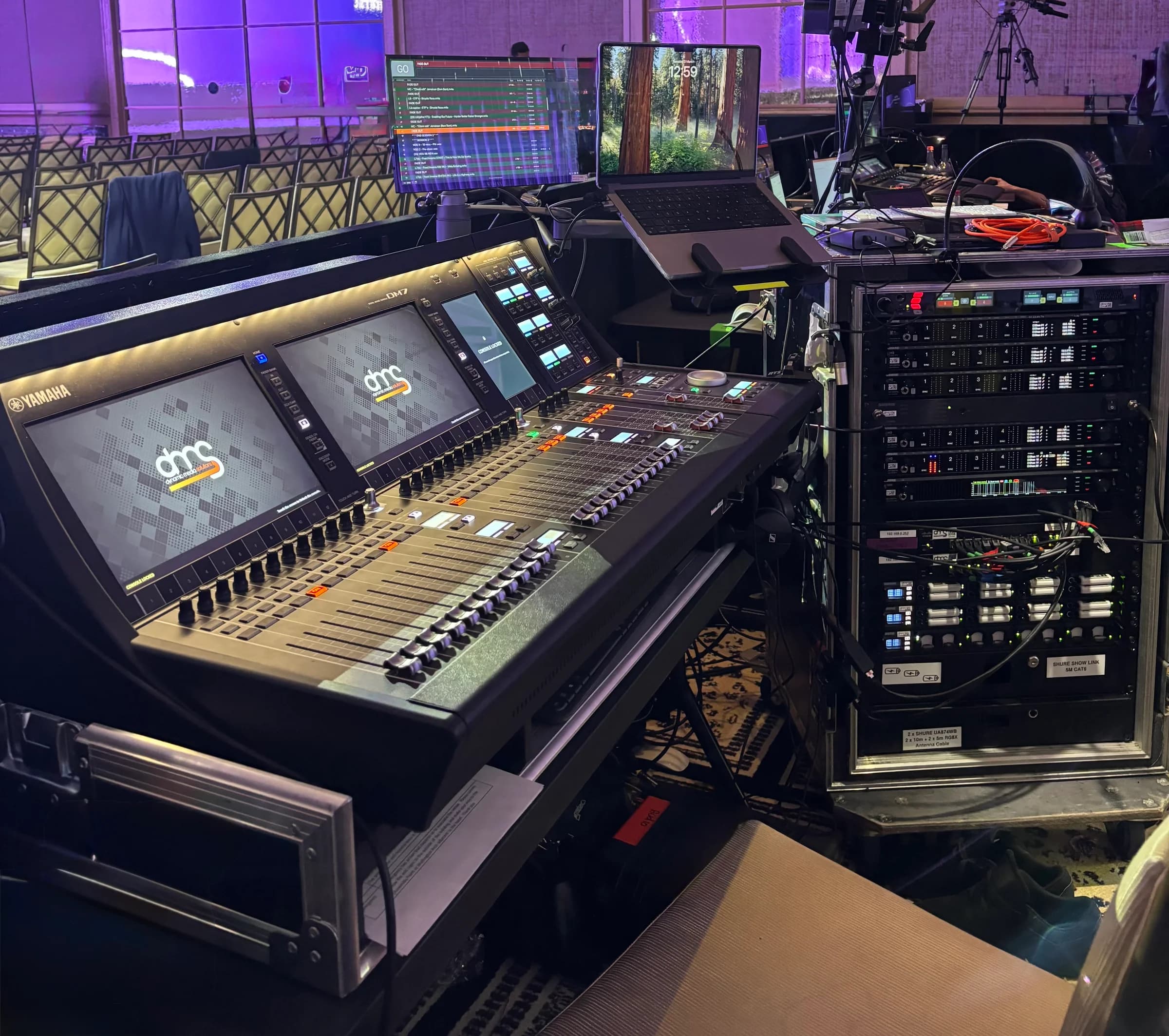 Audio mixing console, equipment racks, a laptop and production monitor beside rows of audience seating