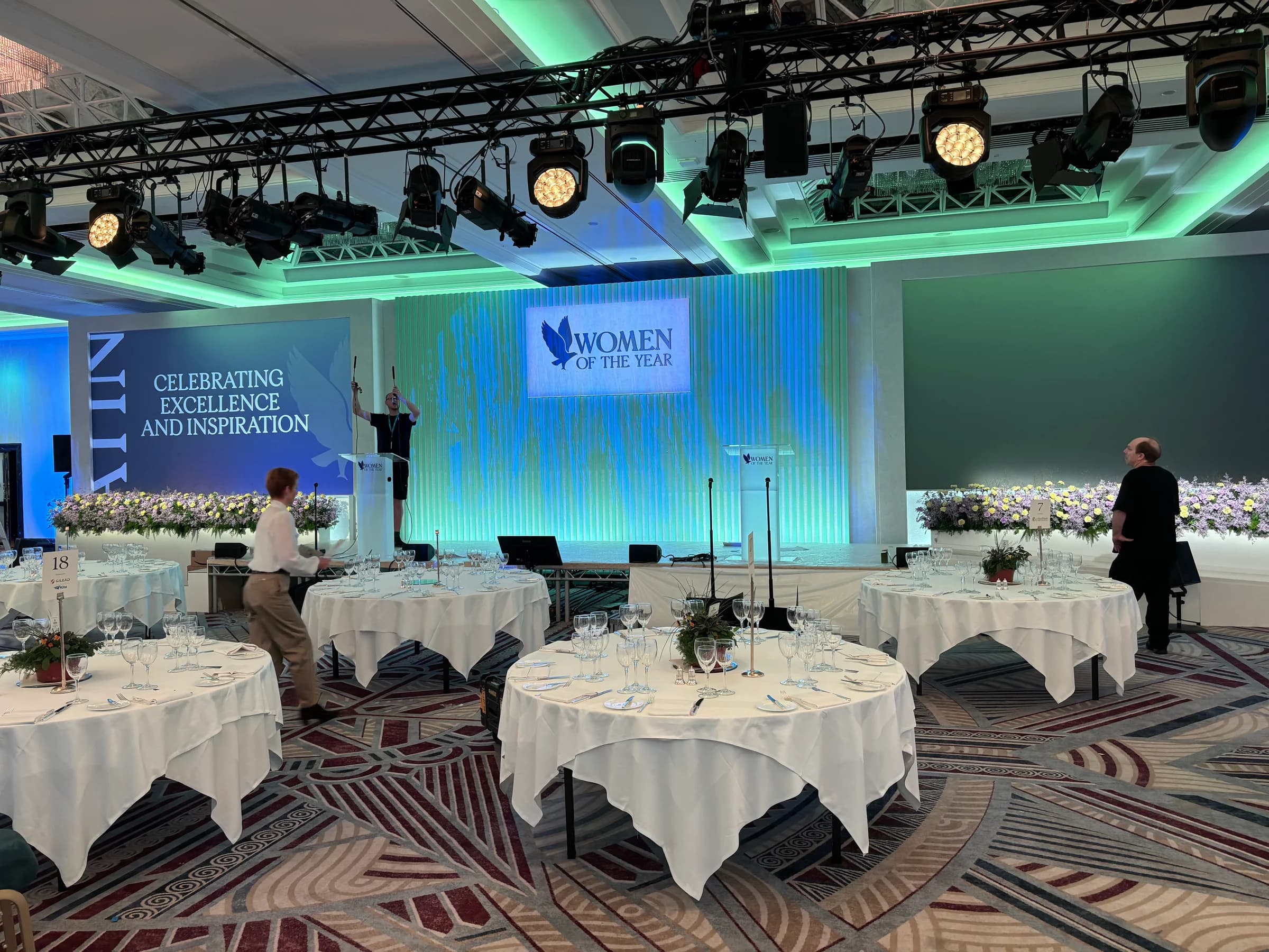 Women of the Year stage with green and blue lighting, overhead lighting truss, clear lecterns and round dining tables
