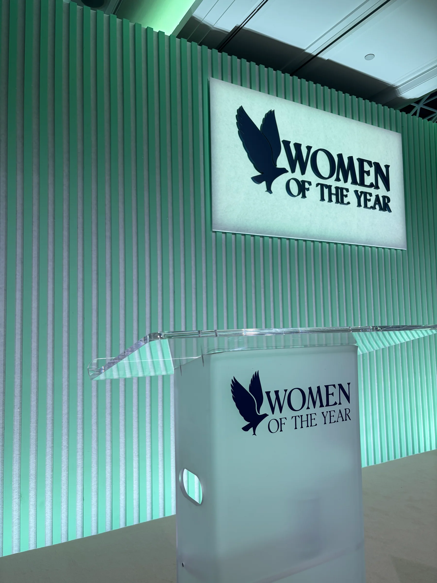 Clear Women of the Year lectern in front of a green-lit backdrop and event sign