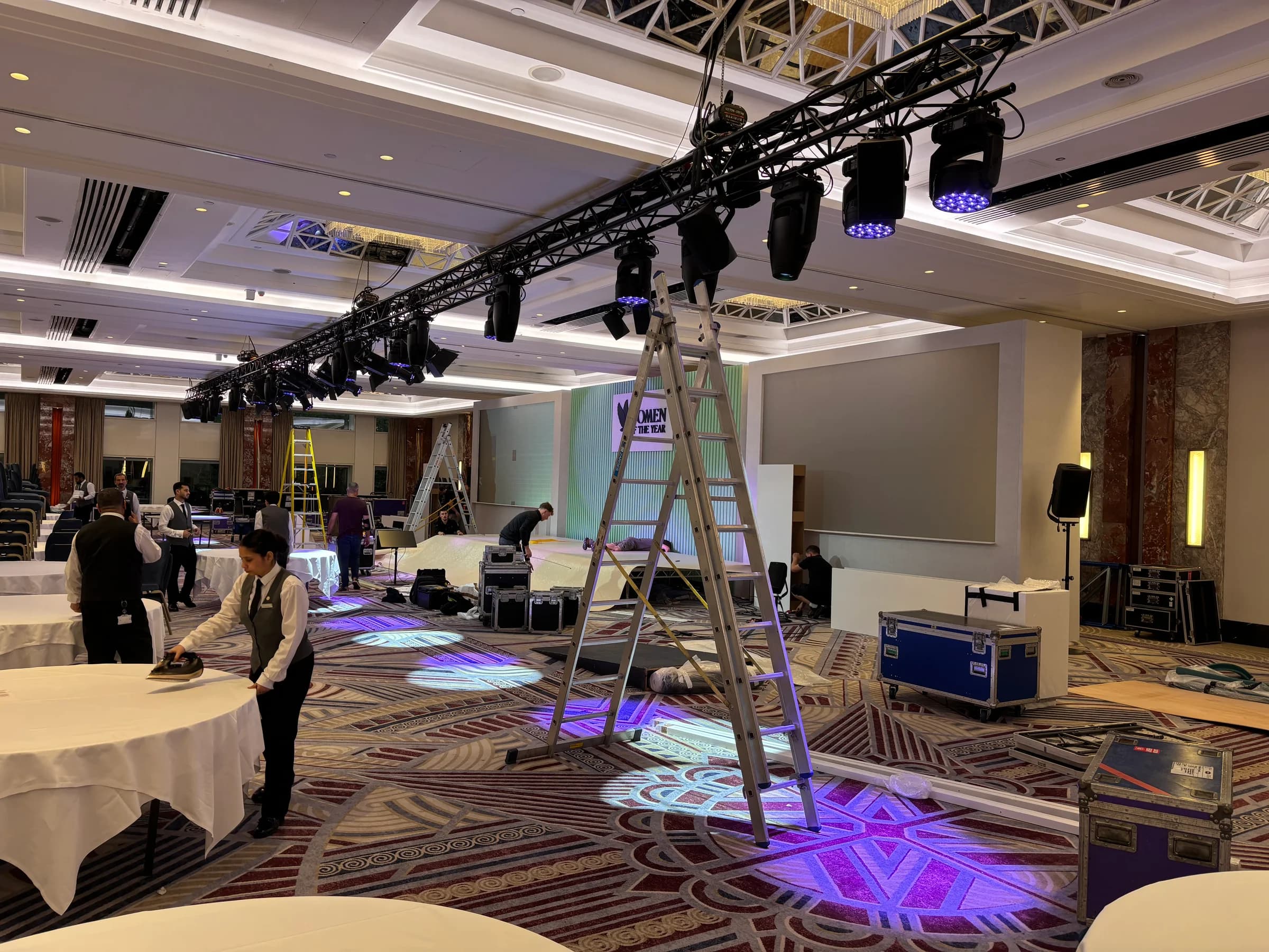 Raised lighting truss above crew preparing the stage and staff dressing dining tables